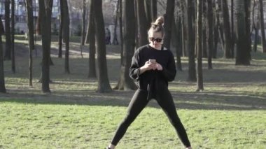 Parktaki fitness antrenmanından önce genç bayan antrenmanı. Sağlıklı genç bir kadın dışarıda ısınıyor. Akıllı telefondaki sosyal ağlardaki yazışmalar dikkatini dağıtıyor. Kadın esneme egzersizi yapıyor.