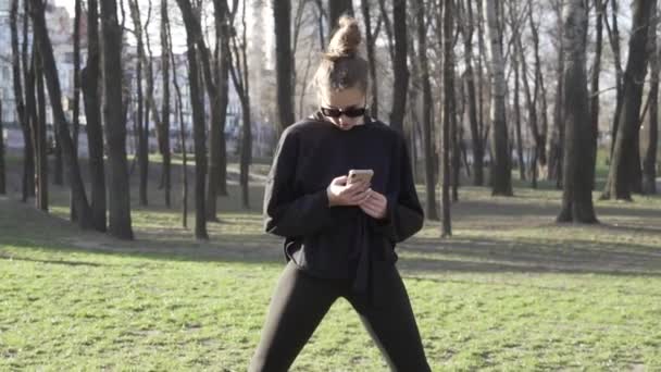 Parktaki fitness antrenmanından önce genç bayan antrenmanı. Sağlıklı genç bir kadın dışarıda ısınıyor. Akıllı telefondaki sosyal ağlardaki yazışmalar dikkatini dağıtıyor. Kadın esneme egzersizi yapıyor. — Stok video