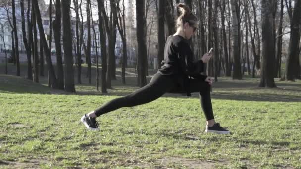 Ung attraktiv kvinna som använder mobiltelefon medan du tränar i parken. Friska unga kvinna värma upp utomhus distraheras av korrespondens i sociala nätverk i en smartphone. hälsosam fitness livsstil — Stockvideo