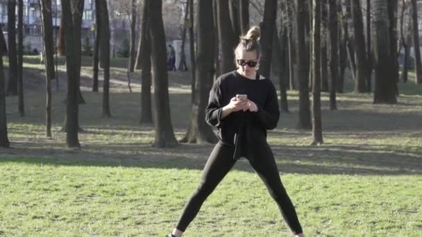 Entrenamiento femenino antes de la sesión de entrenamiento de fitness en el parque. Una mujer joven sana que se calienta al aire libre distraído por la correspondencia en las redes sociales en un teléfono inteligente. mujer que hace ejercicio de estiramiento — Vídeo de stock