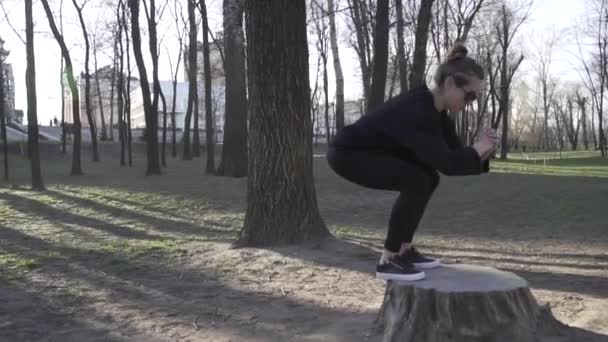 Sportconcept, vetverbranding en gezonde levensstijl. Training en oefeningen voor spieren van benen op de natuur in het park. Een vrouw die uit grijs springt voor een stomp in een park. Meisje atleet hurkt en springt — Stockvideo