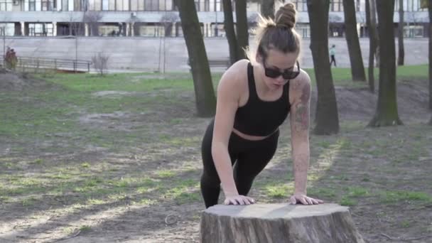 Empurre para cima ou pressione o exercício pela jovem mulher. Jovem fazendo flexões nas madeiras. Atleta fêmea fazendo exercícios para músculos do braço push-ups de uma árvore derrubada em um parque — Vídeo de Stock