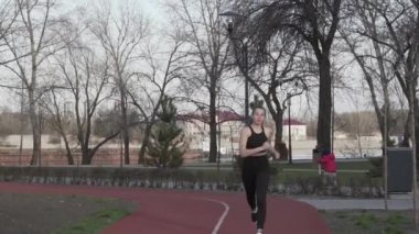 Genç beyaz bir kadın, kırmızı lastik bir yolda parkta koşuyor. Kız sporcu şehirde kardiyo çalışması yapıyor. Hareket hayattır. Şehir parkında koşan sporcu kadın.