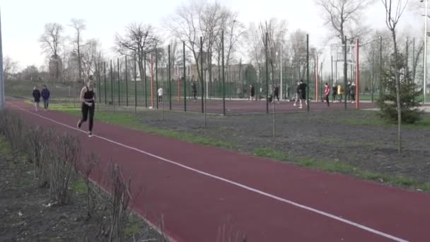 Uma jovem caucasiana corre ao longo de uma pista de borracha vermelha através de um parque. Atleta menina fazendo exercício cardio na cidade. Movimento é vida. Sportswoman jogging em um parque da cidade — Vídeo de Stock