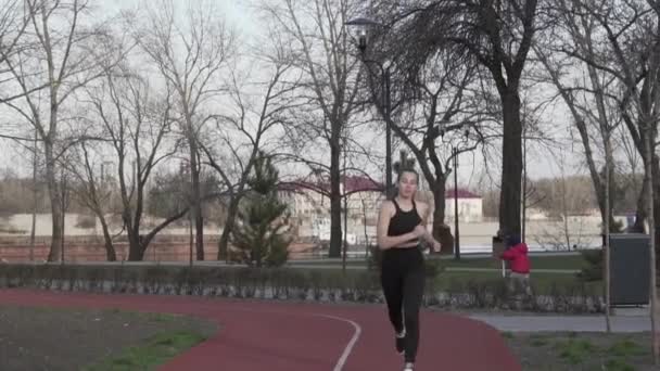 A young Caucasian woman runs along a red rubber track through a park. Girl athlete doing cardio workout in the city. Movement is life. Sportswoman jogging in a city park — Stock Video