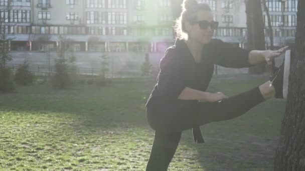 Jonge sportieve vrouw doet stretching oefeningen in de buurt van boom in het park. Meisje rust na het sporten buiten in de natuur. Een atletische vrouw die zich tegen een boom uitstrekt. fit sportvrouw doet warming-up oefeningen — Stockvideo