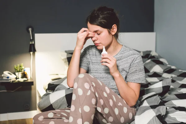 The theme is seasonal cold, runny nose flu virus infection. Young caucasian woman at home bedroom bed uses spray drops of medicine in her nose for snot. Allergic renitis and sinusitis.