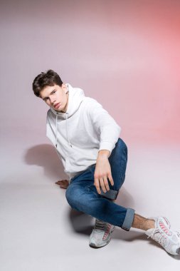 Beyaz eşofman altı, mavi kot pantolon ve beyaz spor ayakkabılar giyen yakışıklı Avrupalı bir erkek model, ticari fotoğraf çekimi için pembe bir arka planda poz veriyor, kameraya bakıyor..