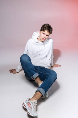 Stüdyoda yerde oturan yakışıklı, şık genç adam. Gündelik kıyafetlerle pembe arka planda otururken poz veren genç bir model. Kot pantolonlu, spor ayakkabılı ve kapüşonlu beyaz ceketli adam..