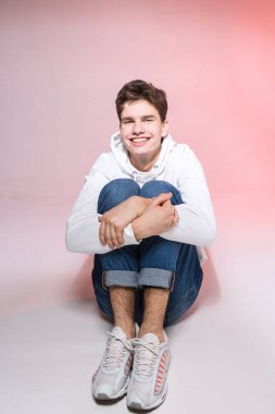 Genç beyaz erkek model stüdyoda yerde otururken poz veriyor. Pembe arka planda şık giyinmiş genç bir çocuk. Kot giyen adam, beyaz başlıklı ve spor ayakkabılı..