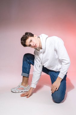 Genç beyaz erkek model stüdyoda yerde otururken poz veriyor. Pembe arka planda şık giyinmiş genç bir çocuk. Kot giyen adam, beyaz başlıklı ve spor ayakkabılı..