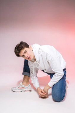 Beyaz eşofman altı, mavi kot pantolon ve beyaz spor ayakkabılar giyen yakışıklı Avrupalı bir erkek model, ticari fotoğraf çekimi için pembe bir arka planda poz veriyor, kameraya bakıyor..