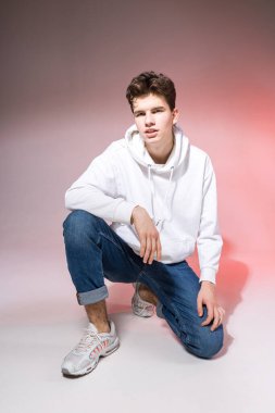 Stüdyoda yerde oturan yakışıklı, şık genç adam. Gündelik kıyafetlerle pembe arka planda otururken poz veren genç bir model. Kot pantolonlu, spor ayakkabılı ve kapüşonlu beyaz ceketli adam..