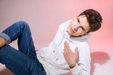Genç beyaz erkek model stüdyoda yerde otururken poz veriyor. Pembe arka planda şık giyinmiş genç bir çocuk. Kot giyen adam, beyaz başlıklı ve spor ayakkabılı..