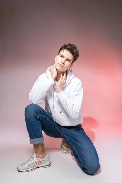 スタジオの床に座っているハンサムな若いスタイリッシュな男 カジュアルな服装でピンクの背景に座っている間にポーズ十代のモデル ジーンズの男スニーカーフード付きの白いジャケット — ストック写真