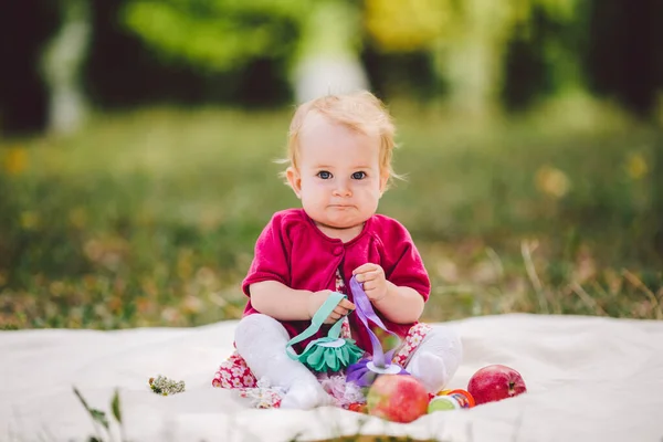 Little baby girl Caucasian ethnicity blond one year old from birth sits on a plaid on green grass in the park. Child at picnic in the autumn forest. Beautiful cute joyful funny baby sitting on blanket