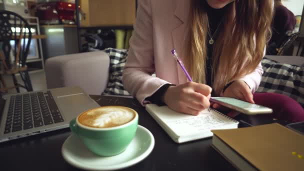 Zamykam kaukaski kobieta ręka na drewniany stół wewnątrz kawiarnia robię notatka w notatnik. Temat niezależny bloger dziennikarz w pracy. Nierozpoznawalna osoba. Technologia telefon i laptop z filiżanką kawy — Wideo stockowe