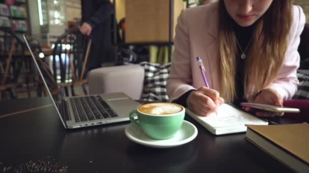 Großaufnahme kaukasische Frau Hand auf Holztisch im Café macht Notizen in Notizbuch. Thema freier Blogger Journalist bei der Arbeit. Bis zur Unkenntlichkeit. Technologie-Telefon und Laptop mit Tasse Kaffee — Stockvideo