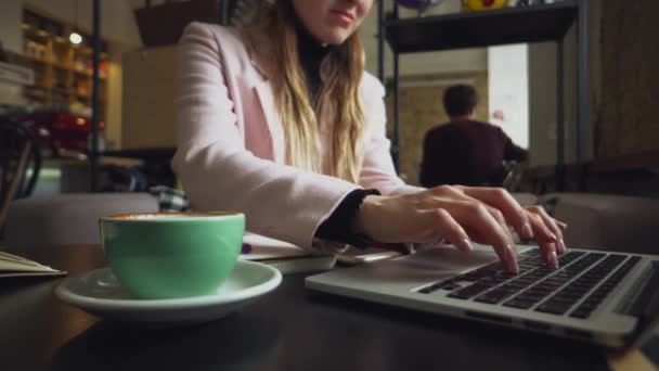 Wanita muda kaukasia yang cantik minum kopi dan mengetik di keyboard di dalam kafe di meja kayu. Topik profesi modern adalah seorang narablog, pekerja lepas, dan penulis — Stok Video