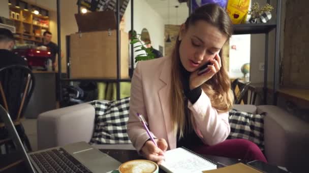 Ładna kobieta w garniturze rozmawiająca przez telefon komórkowy, siedząca w kawiarni, pracująca z laptopem i notatnikiem. Kobieta freelancer mówi na smartfonie i pisanie w notatniku — Wideo stockowe