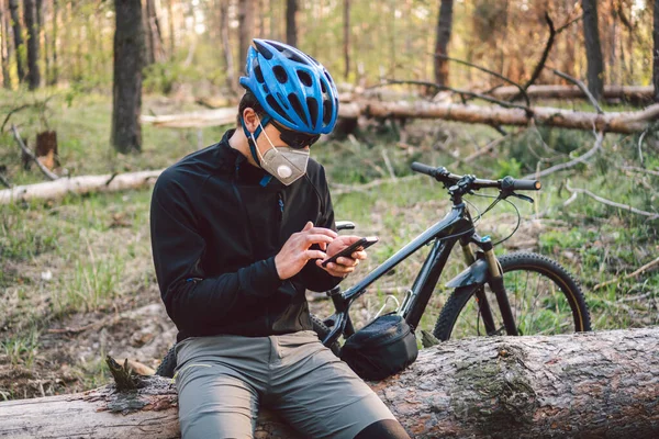 Male cyclist wearing respirator face mask with heavy duty protective filter, sitting in forest and uses phone. safety device for protect health. Cyclist in pollution mask from bushfire smoke haze.
