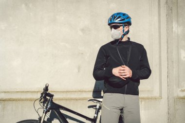 Koruyucu yüz maskesi takmış bir bisikletçi gri beton duvarın arka planında bir bisikletin yanında duruyor. Solunum cihazında tanımlanamayan kurye. Bir salgın sırasında teslimat servisi.