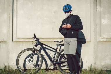 Bisikletli koruyucu maskeli kurye durdu ve telefon uygulamasını izliyordu. Karantinada teslimat servisi. Solunum cihazındaki bir adam cep telefonu kullanıyor. Bisikletli kurye çalışıyor.
