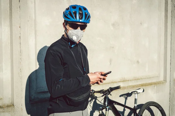 The courier in protective mask on bicycle stopped and was watching the phone application. Delivery service in quarantine. A man in a respirator uses a mobile phone. Bicycle messenger works.
