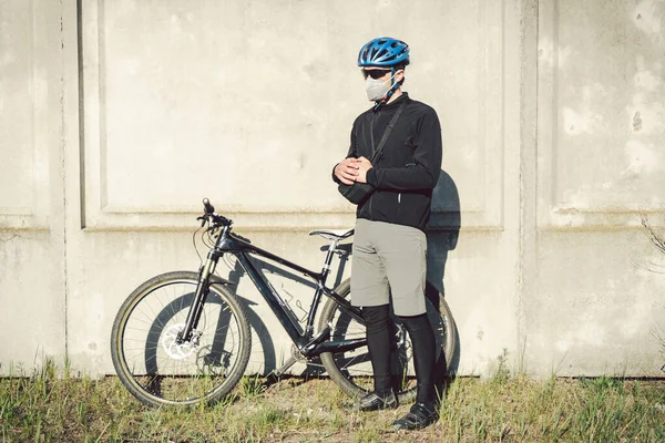 A man cyclist in a protective face mask stands next to a bicycle on a background of gray concrete wall. Unrecognizable Courier in respirator. Delivery service during a pandemic.