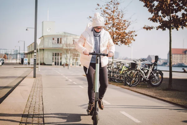 Young woman riding an electric scooter on bike path in copenhagen, modern girl, new generation, electric transport in city, ecology and ecological transport. Woman renting electric skateboard in city.