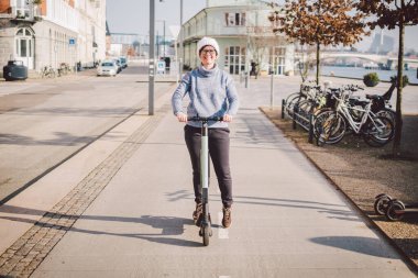 Kopenhag 'da bisiklet yolunda elektrikli scooter süren genç bir kadın, modern kız, yeni nesil, şehirde elektrik nakli, ekoloji ve ekolojik ulaşım. Şehirde elektrikli kaykay kiralayan bir kadın..