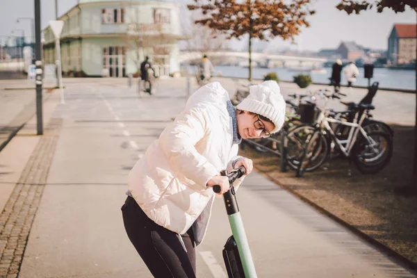 Young woman riding an electric scooter on bike path in copenhagen, modern girl, new generation, electric transport in city, ecology and ecological transport. Woman renting electric skateboard in city.