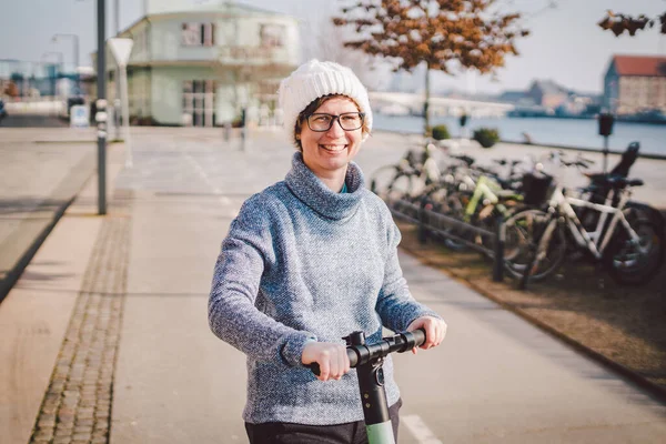 Young woman riding an electric scooter on bike path in copenhagen, modern girl, new generation, electric transport in city, ecology and ecological transport. Woman renting electric skateboard in city.