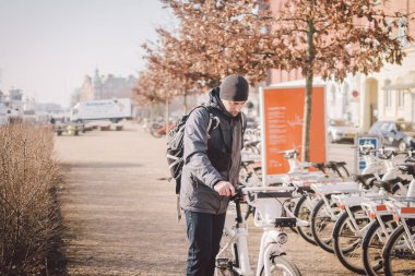 Adam Kopenhag 'da elektrikli beyaz bisiklet kiralıyor. E-Bisiklet Kiralama İstasyonu. Erkek park etmiş şehir bisikletleri, karbondioksit ve yeşil enerjiden tasarruf ediyoruz diyor. Genç adam bisikletini yatırıyor. Kiralık motosikletler.