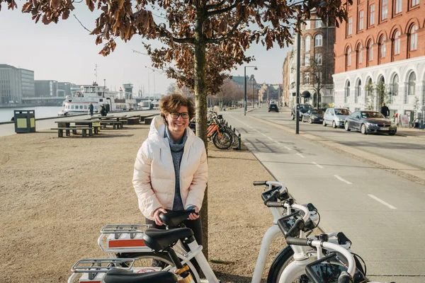 Happy woman renting bicycle at public urban cycle transport station in Copenhagen, Denmark. Young woman taking a shared bicycle. Bike rental. ecological bicycle transport.