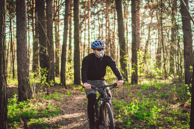 Genç adam ormanda dağ bisikleti sürüyor. Parkta bisiklet süren genç bir atletik adam. Erkek bisikletçi yüz solunum cihazı takıyor, saat 2.5 temalı Coronavirus veya Covid 19. Karantina Sporları.