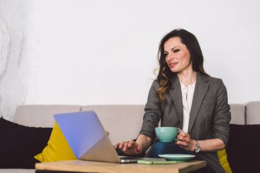 İş yerinde kupa kahveli modern iş kadını. Kafede dizüstü bilgisayarı olan bir iş kadını. Ofis elbiseli orta yaşlı bayana kahve molası. Kahve dükkanında çalışırken sıcak bir şeyler içiyor..