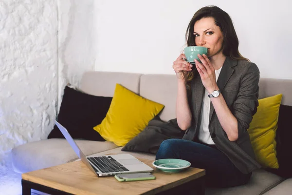 Cheerful skilled middle aged life or business coach enjoying creative process of making new text, sitting at workspace, writing down thoughts, ideas with mug, laptop on desk in coffe shop.