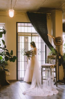 Gelin büyük an için bekliyor. Güzel elbiseli genç gelin elinde çiçeklerle evin penceresinin yanında poz veriyor. Düğün konsepti. Müstakbel karısı düğün gününde sevgili erkeğini bekliyor..