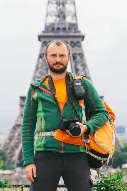 Elinde fotoğraf makinesi ve turuncu sırt çantası olan beyaz bir adam Fransa 'da duruyor. Paris, Eyfel Kulesi' nin arka planında..