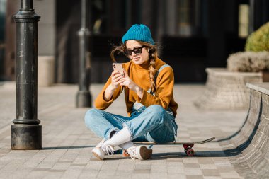 Şapkalı, güneş gözlüklü ve kotlu genç beyaz kadın kaykaycı şehirdeki bir kaykayın yanında telefon kullanıyor. Kentsel bir ortamda aktif gençlik eğlencesi teması.