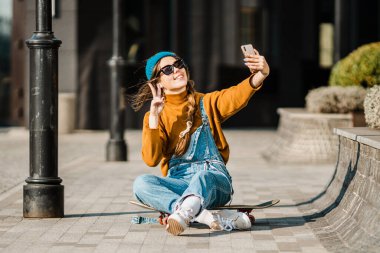 Kaykayın üzerinde oturan ve cep telefonu kullanan bir kız. Açık hava, şehir hayatı. Kaykaycı kız kaykayın üzerinde oturmuş akıllı telefonları kontrol ediyor internet kullanarak müzik dinliyor ve fotoğraf çekiyor..