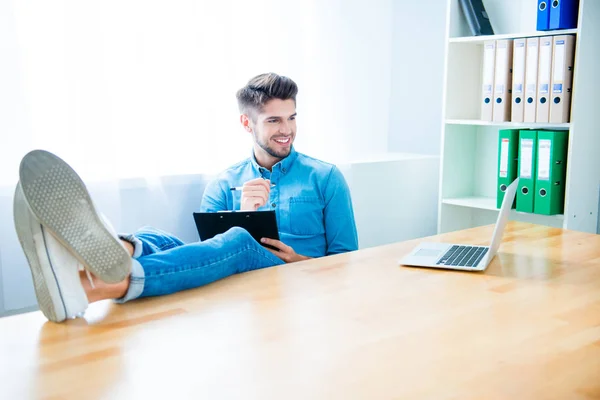 Junger Manager mit Laptop, der Beine auf dem Tisch hält und Notizen macht — Stockfoto