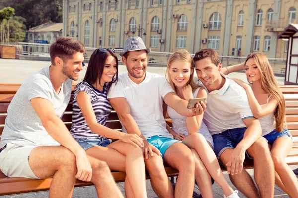 Šest happy radostné přátelé sedí na lavičce a výrobu fotografií na sm — Stock fotografie