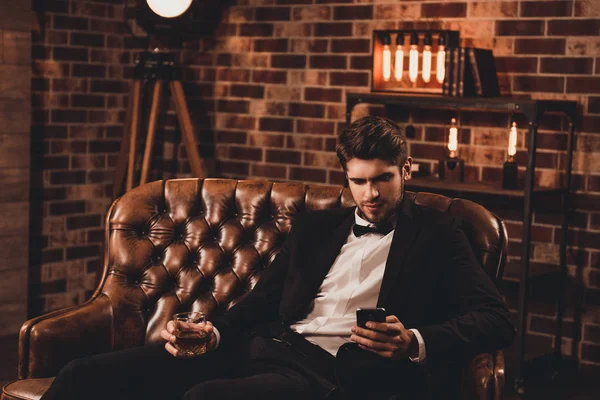 Young man in suit sitting on couch with glass of cognac and typi
