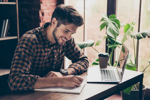 Sonriente estudiante guapo trabajando con el ordenador portátil y tomando notas — Foto de Stock