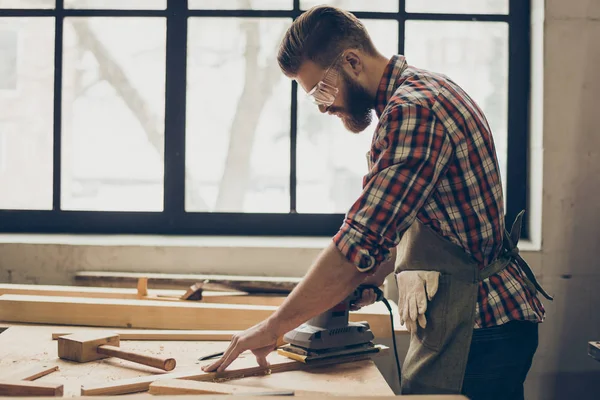 Side view of young bearded craftsman with sander grinding a wood — Stock Photo, Image