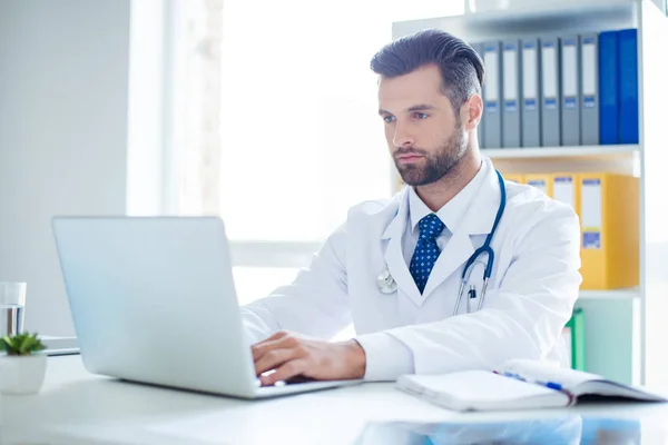 Розумний успішний лікар сидить у своєму кабінеті і використовує ноутбук — стокове фото