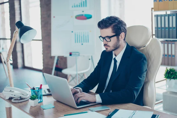 Успешный предприниматель в черной формальной одежде с галстуком сидит в своем современном офисе и использует ноутбук для своей работы — стоковое фото