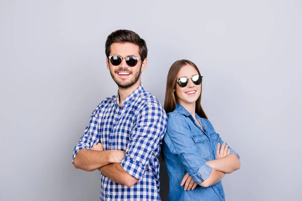 Due giovani amanti allegri guardano nella fotocamera, indossano occhiali da sole alla moda e sorridono, in camicie casual, con le mani incrociate sullo sfondo puro — Foto Stock